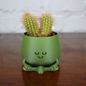 Puede incluir: Una maceta de cer&aacute;mica verde con forma de figura meditando con una planta de cactus dentro. La maceta tiene una cara sonriente con los ojos cerrados.