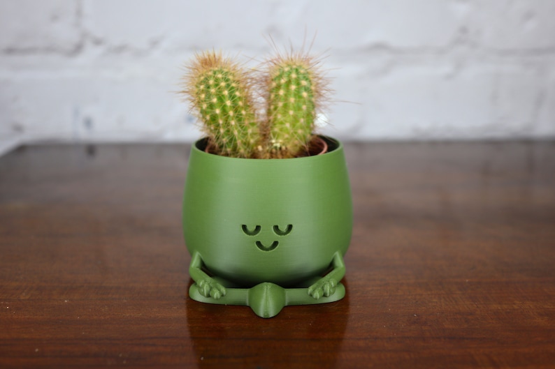 Puede incluir: Una maceta de cer&aacute;mica verde con forma de figura meditando con una planta de cactus dentro. La maceta tiene una cara sonriente con los ojos cerrados.