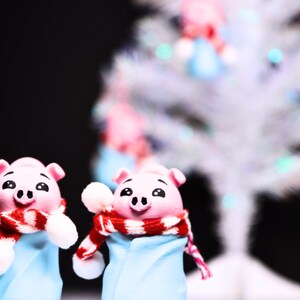 May include: Two pink pig figurines wearing blue and white winter outfits with red and white scarves. The figurines are standing in front of a white, sparkly Christmas tree.
