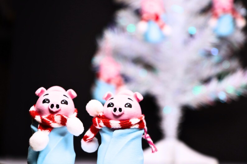 May include: Two pink pig figurines wearing blue and white winter outfits with red and white scarves. The figurines are standing in front of a white, sparkly Christmas tree.