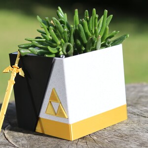 May include: A white and black cube-shaped planter with a gold trim and a gold triangular symbol. A small gold sword is leaning against the planter. A green succulent plant is growing inside the planter.