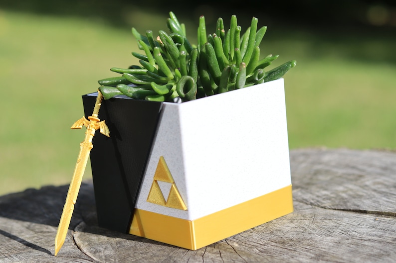 May include: A white and black cube-shaped planter with a gold trim and a gold triangular symbol. A small gold sword is leaning against the planter. A green succulent plant is growing inside the planter.