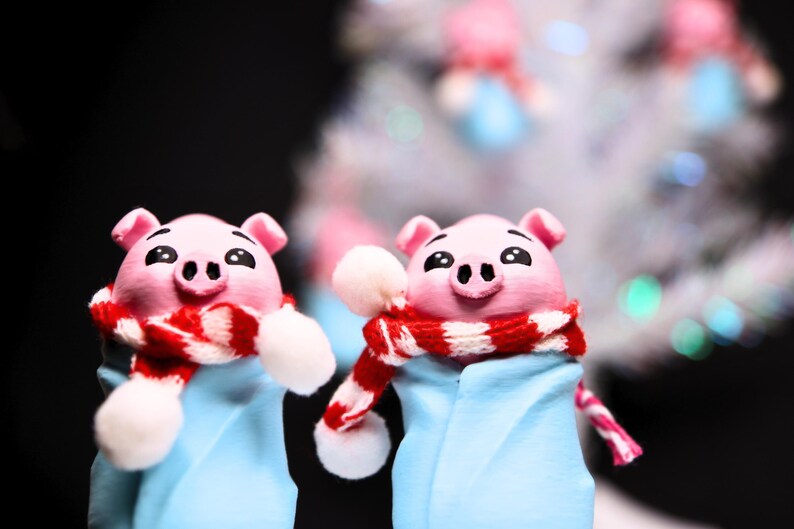 May include: Two pink pig figurines wearing blue and white scarves with red and white stripes. The scarves have white pom-poms on the ends. The figurines are in front of a blurry white background.