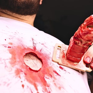 May include: A white shirt with a large, bloody hole in the center. A red, plastic alien creature is being held by a hand, with the creature's head sticking out of the hole in the shirt.