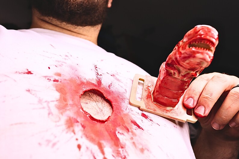 May include: A white shirt with a large, bloody hole in the center. A red, plastic alien creature is being held by a hand, with the creature's head sticking out of the hole in the shirt.