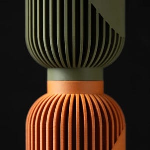 Peut inclure: Objet d&eacute;coratif en deux parties avec un design nervur&eacute;. La partie sup&eacute;rieure est vert olive avec un accent g&eacute;om&eacute;trique, et le bas est orange. L'objet a une forme cylindrique avec des rainures verticales.