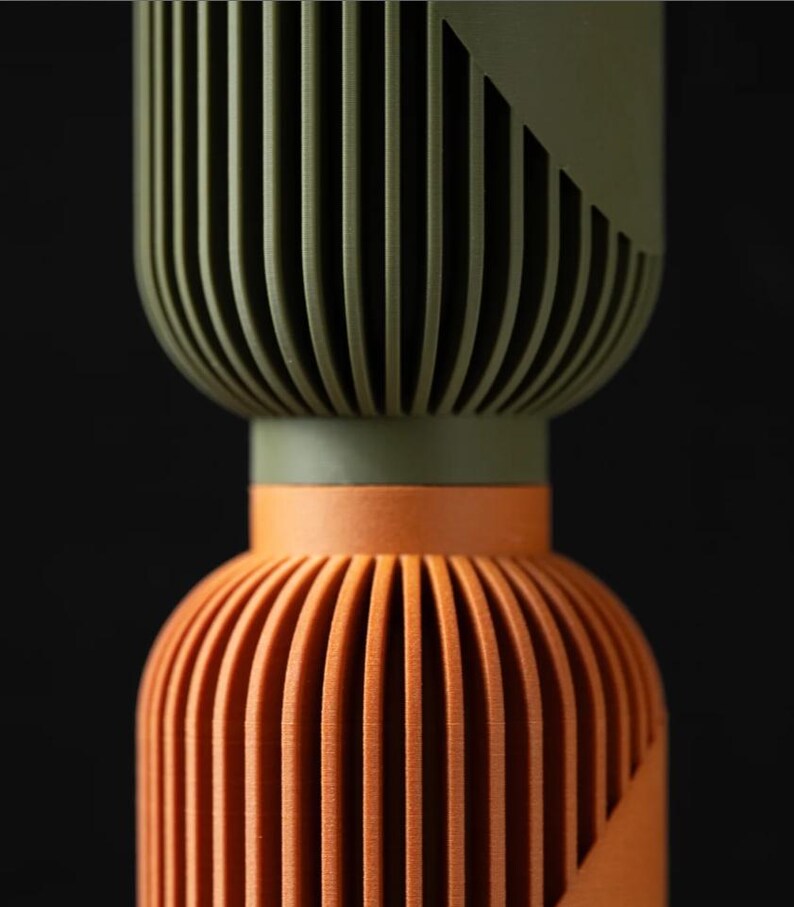Peut inclure: Objet d&eacute;coratif en deux parties avec un design nervur&eacute;. La partie sup&eacute;rieure est vert olive avec un accent g&eacute;om&eacute;trique, et le bas est orange. L'objet a une forme cylindrique avec des rainures verticales.