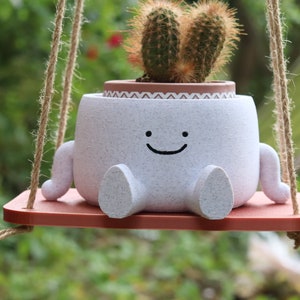 May include: A white ceramic planter shaped like a smiling face with small arms and legs, holding a cactus plant. The planter is hanging from a brown rope swing.
