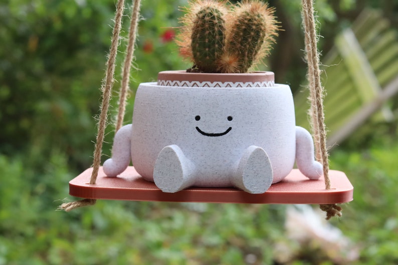 May include: A white ceramic planter shaped like a smiling face with small arms and legs, holding a cactus plant. The planter is hanging from a brown rope swing.