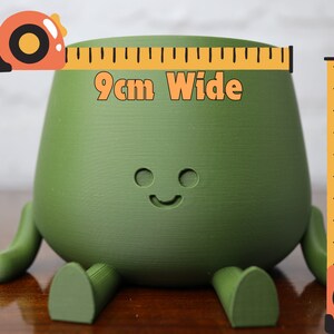Puede incluir: Una maceta de cer&aacute;mica verde con forma de personaje de dibujos animados con piernas y una cara sonriente. La maceta mide 9 cm de ancho y 9 cm de profundidad.