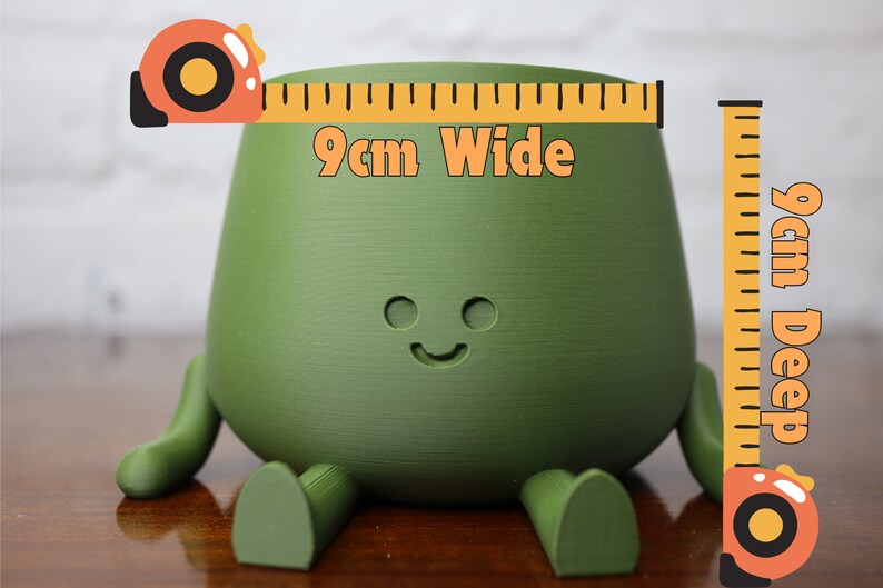Puede incluir: Una maceta de cer&aacute;mica verde con forma de personaje de dibujos animados con piernas y una cara sonriente. La maceta mide 9 cm de ancho y 9 cm de profundidad.