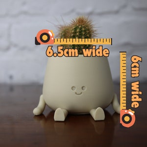 Puede incluir: Una maceta de cer&aacute;mica beige claro con una cara sonriente y patas, que sostiene una peque&ntilde;a planta de cactus. La maceta mide 6,5 cm de ancho y 6 cm de alto.
