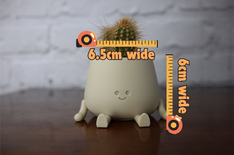 Puede incluir: Una maceta de cer&aacute;mica beige claro con una cara sonriente y patas, que sostiene una peque&ntilde;a planta de cactus. La maceta mide 6,5 cm de ancho y 6 cm de alto.