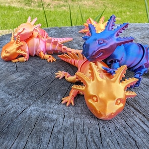 May include: Four axolotl figurines, each with a different iridescent color. The axolotls are posed in a playful manner on a wooden surface. The colors are pink, orange, blue, and purple.