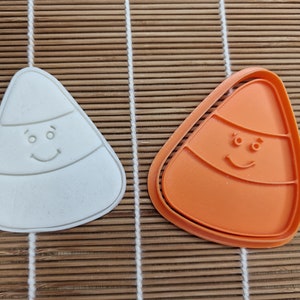 May include: Two candy corn-shaped cookie cutters. One is white, the other is orange. Both have a smiling face design. The cutters are on a bamboo mat. Great for Halloween baking.