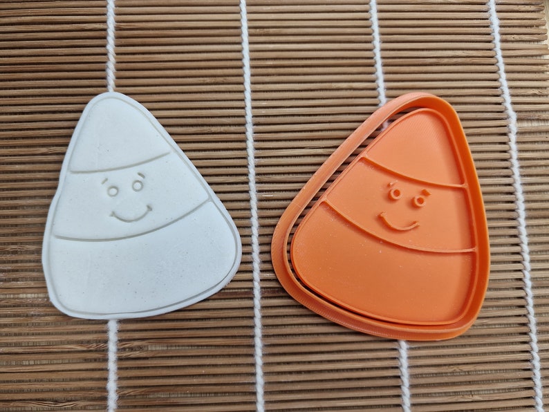 May include: Two candy corn-shaped cookie cutters. One is white, the other is orange. Both have a smiling face design. The cutters are on a bamboo mat. Great for Halloween baking.
