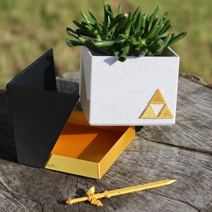 May include: A white planter with a gold triangular symbol, a black and orange box, and a gold sword. The planter has a green succulent plant inside. The box is open and the sword is lying on a wooden surface.