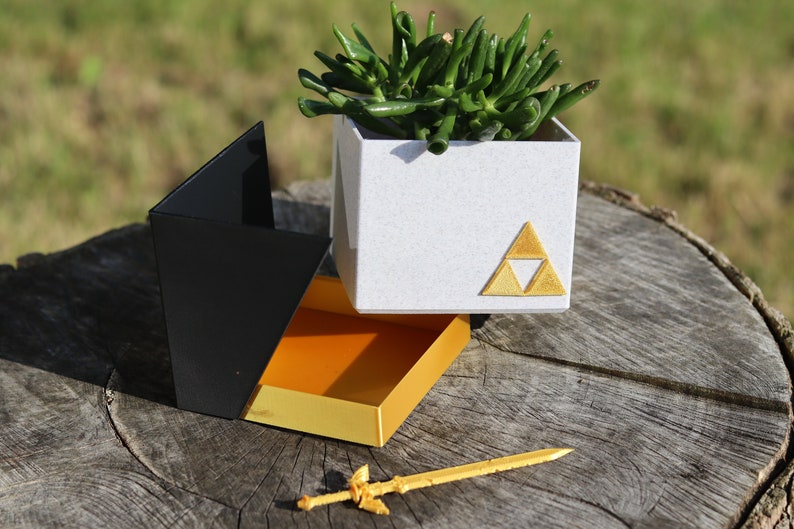 May include: A white planter with a gold triangular symbol, a black and orange box, and a gold sword. The planter has a green succulent plant inside. The box is open and the sword is lying on a wooden surface.