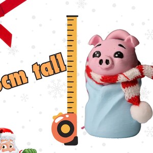May include: A small pink pig wearing a red and white scarf and a blue blanket. The pig is 1.5 cm tall and is standing next to a measuring tape.