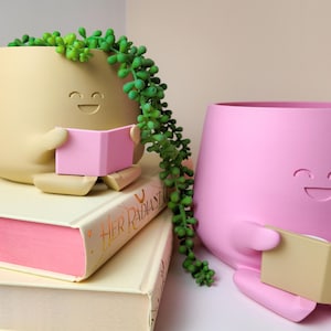 Puede incluir: Dos macetas de cer&aacute;mica con forma de caras sonrientes, ambas sosteniendo libros abiertos. La maceta de la izquierda es beige con un libro rosa y est&aacute; sentada sobre dos libros. La maceta de la derecha es rosa con un libro beige.