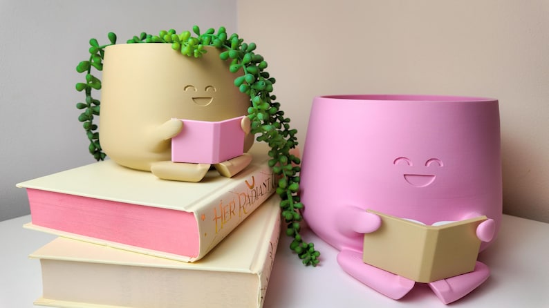 Puede incluir: Dos macetas de cer&aacute;mica con forma de caras sonrientes, ambas sosteniendo libros abiertos. La maceta de la izquierda es beige con un libro rosa y est&aacute; sentada sobre dos libros. La maceta de la derecha es rosa con un libro beige.