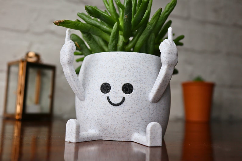May include: White ceramic planter shaped like a person with arms raised and a smiling face. The planter has a speckled texture and is holding a green succulent plant.