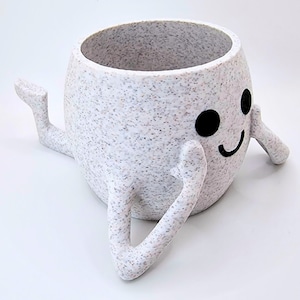 May include: White ceramic planter with a speckled finish shaped like a smiling face with two arms. The planter is sitting on a white surface.