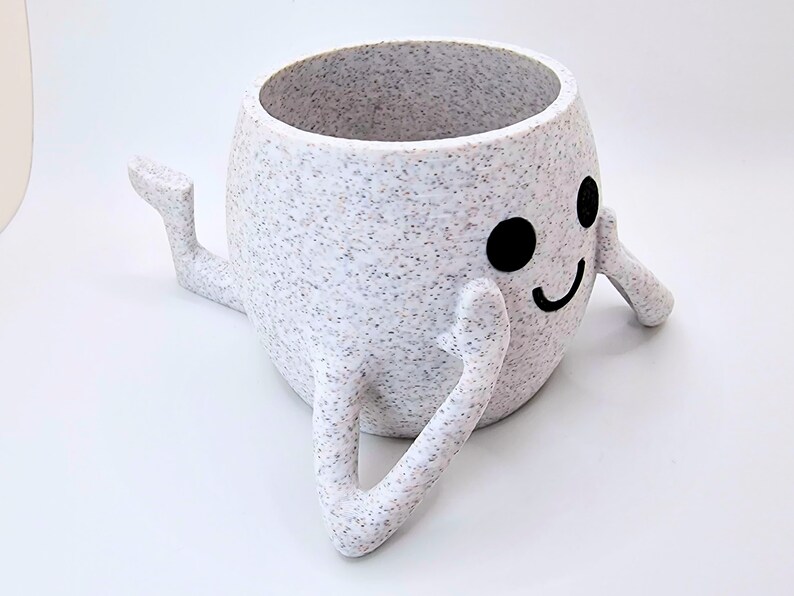 May include: White ceramic planter with a speckled finish shaped like a smiling face with two arms. The planter is sitting on a white surface.
