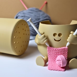 May include: A yellow ceramic yarn bowl with a smiling face and two arms holding knitting needles. The bowl is holding a pink knitted scarf and a ball of pink yarn.