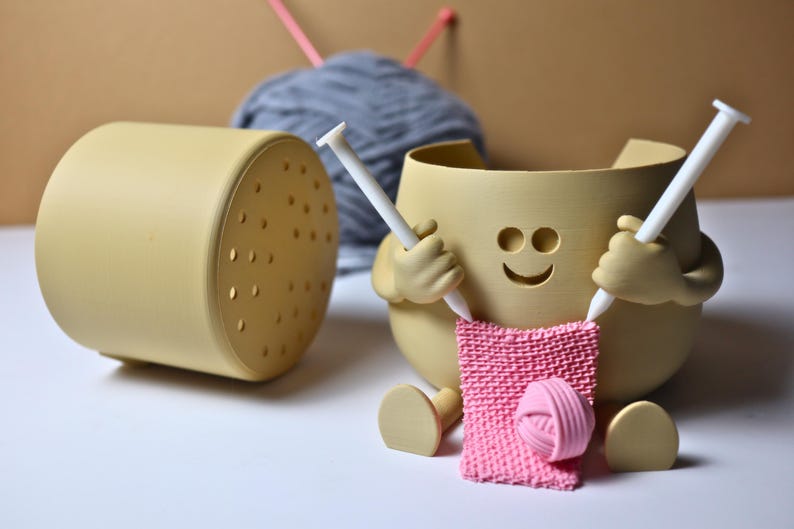 May include: A yellow ceramic yarn bowl with a smiling face and two arms holding knitting needles. The bowl is holding a pink knitted scarf and a ball of pink yarn.