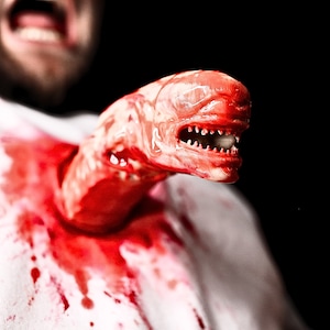 May include: A close-up of a red alien creature bursting out of a white shirt, with blood splattered on the fabric. The alien has sharp teeth and a menacing expression.