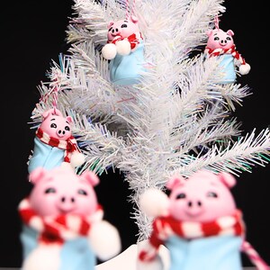 May include: Five pink pig ornaments with blue and red scarves hanging on a white artificial Christmas tree.