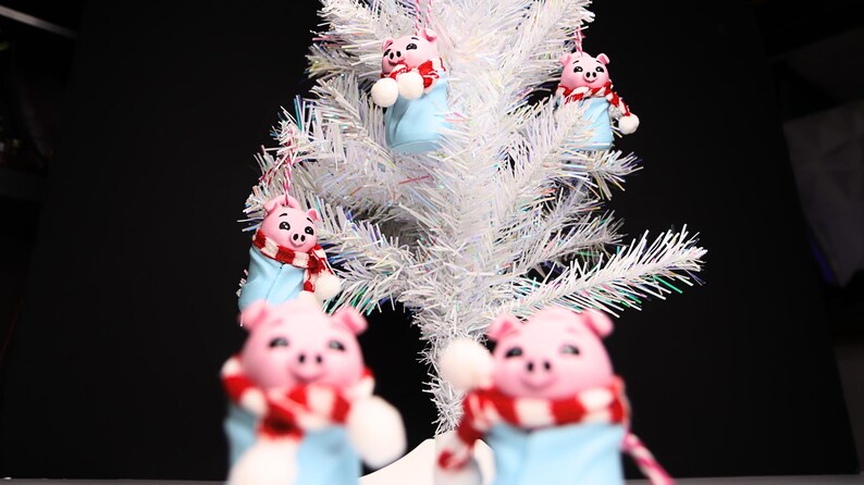 May include: Five pink pig ornaments with blue and red scarves hanging on a white artificial Christmas tree.