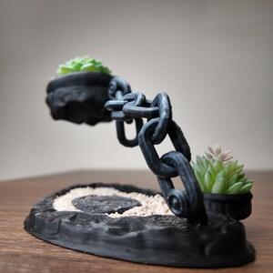 May include: A black, 3D-printed succulent planter with a chain design. The planter features two small pots holding green and pink succulents, connected by a chain. The base has a textured, wood-like appearance with a sand-filled centre.