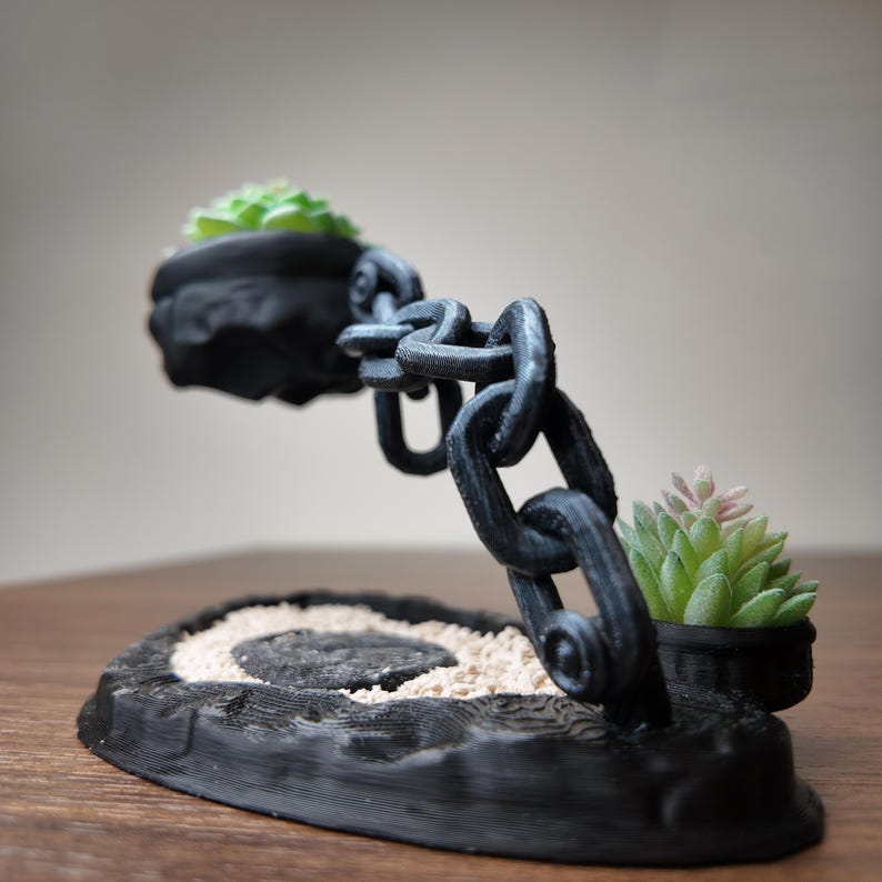 May include: A black, 3D-printed succulent planter with a chain design. The planter features two small pots holding green and pink succulents, connected by a chain. The base has a textured, wood-like appearance with a sand-filled centre.