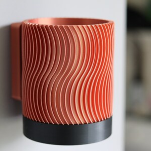 May include: A copper-colored wall-mounted cup holder with a wavy, textured design. The holder has a black base.