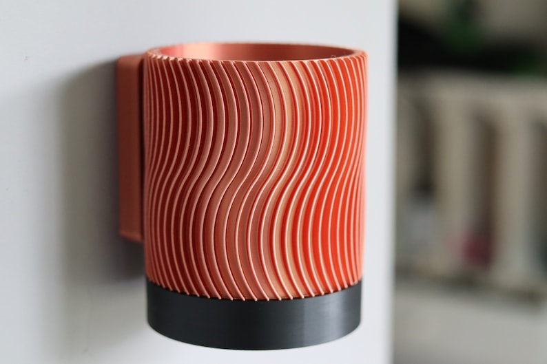 May include: A copper-colored wall-mounted cup holder with a wavy, textured design. The holder has a black base.