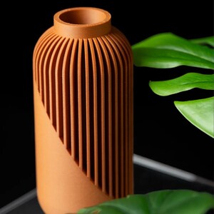 Peut inclure: Un vase de couleur terre cuite avec un corps cylindrique et un col &eacute;troit. Le vase pr&eacute;sente des d&eacute;tails verticaux et stri&eacute;s sur la partie sup&eacute;rieure, cr&eacute;ant un effet textur&eacute;. Le design comprend une coupe diagonale. Le fond est noir.