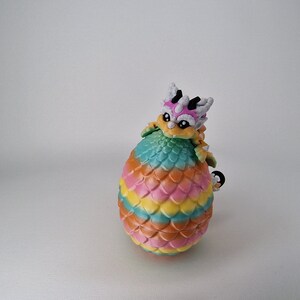 Puede incluir: Una figurita de drag&oacute;n colorida con una c&aacute;scara de huevo arco&iacute;ris. El drag&oacute;n es blanco, rosa y amarillo con ojos negros. La c&aacute;scara del huevo es un degradado de naranja, amarillo, rosa, verde y azul.