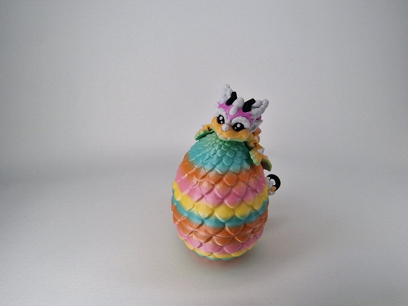 Puede incluir: Una figurita de drag&oacute;n colorida con una c&aacute;scara de huevo arco&iacute;ris. El drag&oacute;n es blanco, rosa y amarillo con ojos negros. La c&aacute;scara del huevo es un degradado de naranja, amarillo, rosa, verde y azul.