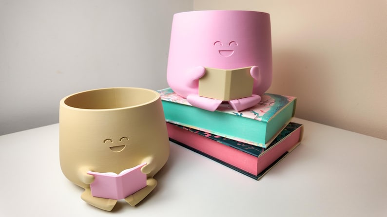 Puede incluir: Dos macetas de cer&aacute;mica con forma de caras sonrientes, cada una sosteniendo un libro. La maceta de la izquierda es beige y la maceta de la derecha es rosa. Las macetas est&aacute;n sobre una pila de tres libros.