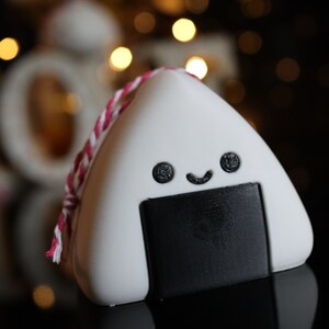 May include: A white and black onigiri shaped ornament with a red and white string. The onigiri has a smiling face with two black eyes and a black mouth.