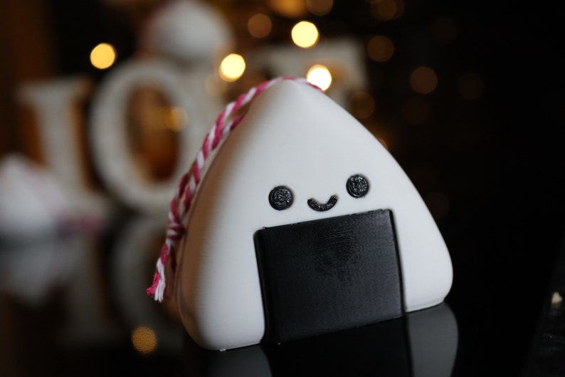 May include: A white and black onigiri shaped ornament with a red and white string. The onigiri has a smiling face with two black eyes and a black mouth.