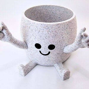 May include: White ceramic planter with a speckled finish shaped like a smiling cartoon character with arms and legs. The planter has black eyes and a black smile.