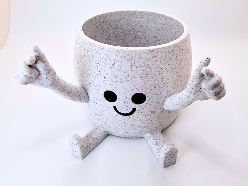 May include: White ceramic planter with a speckled finish shaped like a smiling cartoon character with arms and legs. The planter has black eyes and a black smile.