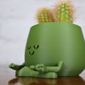Puede incluir: Una maceta de cer&aacute;mica verde con forma de figura meditando con una cara sonriente. La maceta tiene dos peque&ntilde;os cactus que crecen en la parte superior.