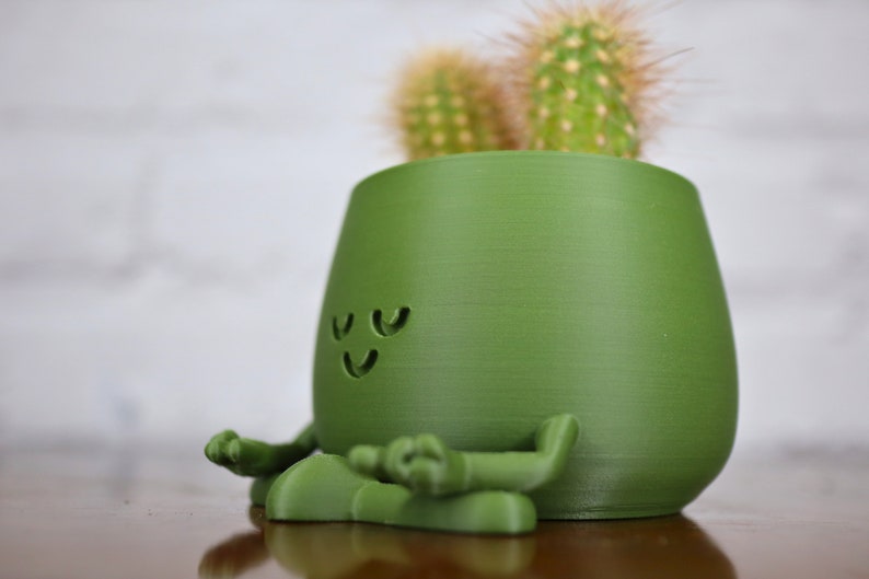 Puede incluir: Una maceta de cer&aacute;mica verde con forma de figura meditando con una cara sonriente. La maceta tiene dos peque&ntilde;os cactus que crecen en la parte superior.