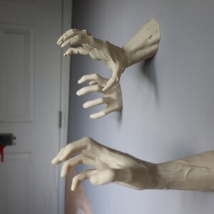 May include: Three realistic-looking plaster hands reaching out from a wall. The hands are painted a pale beige color and appear to be coming out of the wall.