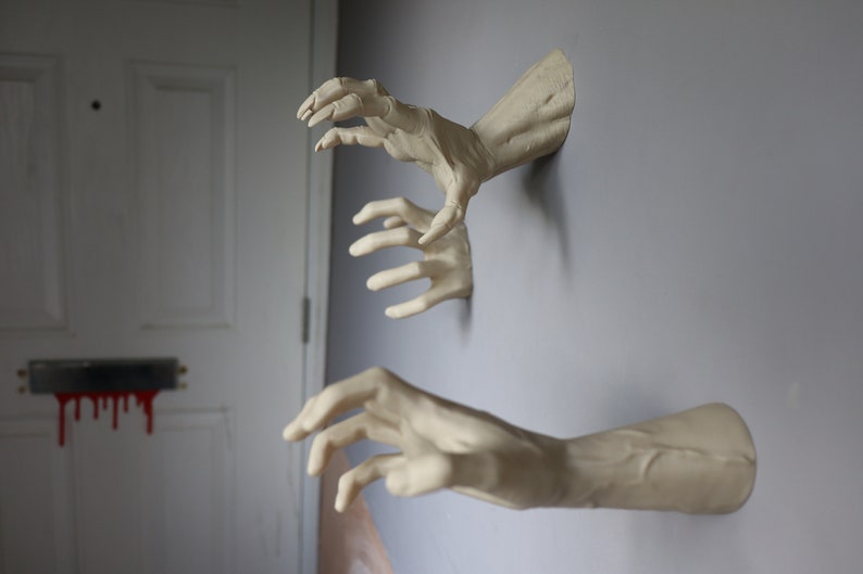 May include: Three realistic-looking plaster hands reaching out from a wall. The hands are painted a pale beige color and appear to be coming out of the wall.