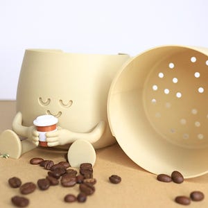 May include: A beige ceramic coffee cup holder with a smiling face and two arms holding a small brown coffee cup. The holder has a removable beige lid with holes for drainage. The holder is sitting on a brown surface with coffee beans scattered around it.